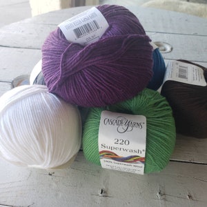 May include: Four balls of yarn in different colors: white, purple, green, and brown. The green ball has a label that reads "Cascade Yarns 220 Superwash 100% Superwash Wool 100g (3.5 oz) / 220 yds (200 m) 10 - 2.75 mm - 11 - 3.25 mm"