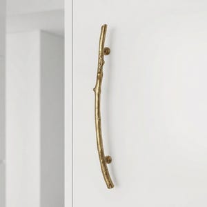 May include: A gold-colored, branch-shaped door pull. The handle is mounted on a white surface. The door pull has a natural, organic design, with a curved shape and small, round knobs at each end.