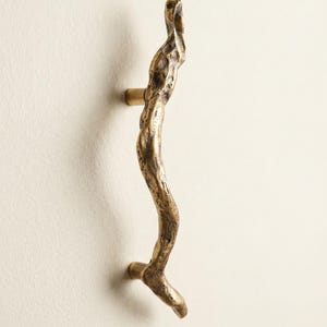 Peut inclure: Une poignée de porte en bronze avec un design texturé et organique. La poignée est incurvée dans une forme naturelle, avec une surface rugueuse et inégale. La poignée est fixée sur un mur blanc, mettant en valeur sa forme artistique unique.