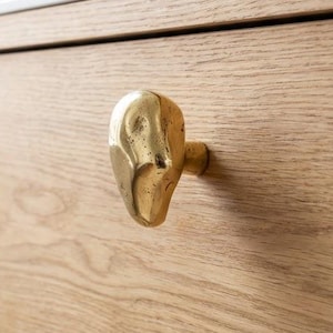 Puede incluir: Primer plano de un mueble de madera con un tirador dorado de forma abstracta. El mueble tiene un acabado de veta de madera clara. El tirador está montado en el cajón superior.