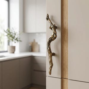 Könnte beinhalten: Ein bronzefarbener Schrankgriff in Form eines Astes. Die dekorative Hardware ist an einer hellbeigen Schranktür mit einer natürlichen Holzverkleidung angebracht. Die Küchenumgebung umfasst ein Fenster, eine Arbeitsplatte und eine Topfpflanze.