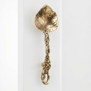 May include: A brass door handle shaped like a leaf. The handle has a detailed leaf design and a stem with grape-like clusters. It is mounted on a white door. The handle is made of brass.