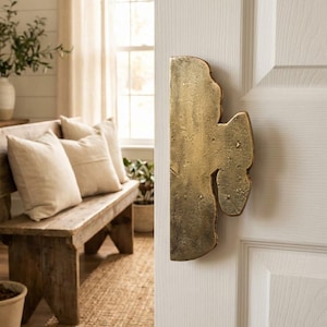 May include: A large, textured brass door pull is mounted on a white door. The door pull has an organic, asymmetrical shape with a warm, golden hue. The background shows a rustic wooden bench with pillows and a jute rug.
