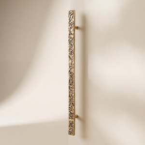 May include: A long, vertical cabinet handle in a metallic gold finish. The handle features a highly textured, irregular, and crinkled surface pattern throughout its length, attached to a flat surface by two small, cylindrical mounting posts.