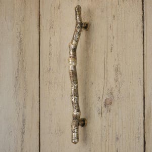 May include: A vertical door handle crafted from metal with a textured, uneven surface that mimics the appearance of a natural tree branch. The handle features a metallic gold and bronze finish and is secured by two circular mounting points.