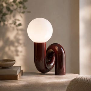 May include: A modern table lamp with a deep red, curved base and a spherical, white light. The lamp's design is minimalist and artistic, suitable for a living room or bedroom. The lamp's height is approximately 46 cm.