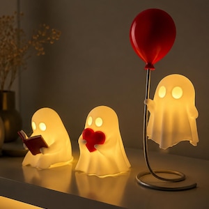 Op de afbeelding: Drie gloeiende spookvormige lampen. Eén leest een boek, een andere houdt een rood hart vast en de derde een rode ballon. De lampen zijn wit met geel licht, wat een grillige en charmante sfeer creëert.