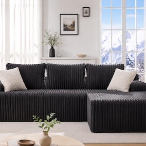 May include: A dark gray corduroy sectional sofa with two cream-colored throw pillows. The sofa has a matching ottoman and is placed in front of a window with a mountain view. The room is decorated with a plant, artwork, and a vase.