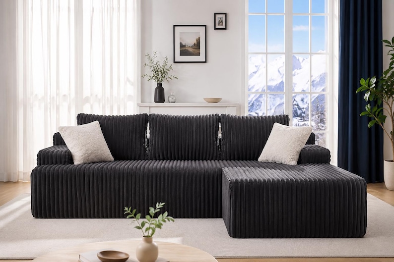 May include: A dark gray corduroy sectional sofa with two cream-colored throw pillows. The sofa has a matching ottoman and is placed in front of a window with a mountain view. The room is decorated with a plant, artwork, and a vase.