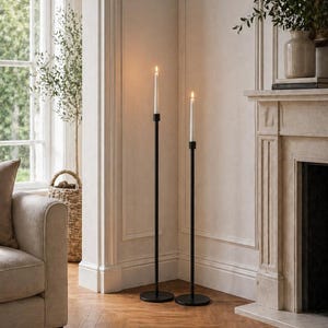 May include: Two black metal floor-standing candle holders, each holding a lit white candle. The holders have a minimalist design and circular bases. The room features a fireplace, window, and wooden flooring, creating a warm atmosphere.
