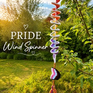 Könnte beinhalten: Ein gehäkeltes Windspiel in Spiralform mit Regenbogenfarben und weißen Rändern. Es enthält einen herzförmigen Anhänger in Pride-Flaggenfarben, Holzperlen und eine mehrfarbige Garnquaste. Der Text PRIDE Wind Spinner ist deutlich zu sehen.