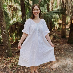 May include: A white, short-sleeved dress with a scoop neckline. The dress is made of a lightweight fabric and has a loose, flowing fit. The person is wearing brown shoes. The background is a lush green forest.