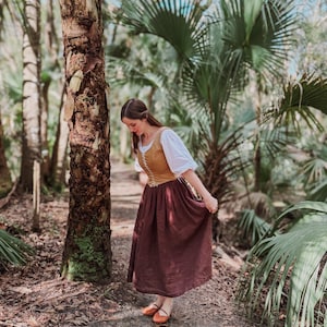May include: A person in a historical costume in a forest setting. The outfit consists of a tan bodice, a white blouse, and a long, dark brown skirt. Brown shoes are worn. The background includes palm trees and other greenery, with sunlight filtering through the trees.