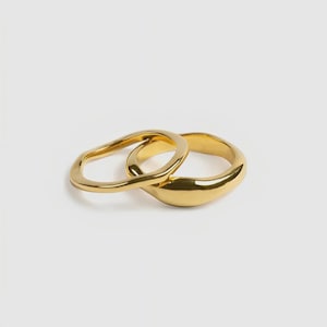 May include: Two gold rings, one with a smooth, rounded band and the other with an irregular, organic shape. The rings are intertwined, creating a unique design. The jewellery is set against a plain white background.
