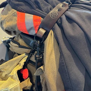 May include: A dark brown leather strap with a circular embossed emblem and black metal hardware, used to secure heavy-duty charcoal and tan protective gear featuring bright orange reflective strips and a yellow mesh pouch with a red accent.