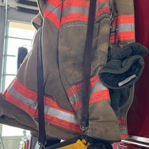 May include: A tan firefighter turnout coat with horizontal reflective orange and silver stripes, featuring a pair of dark grey gloves tucked into the side and a yellow Surei flashlight with a black handle hanging from a long brown leather strap.