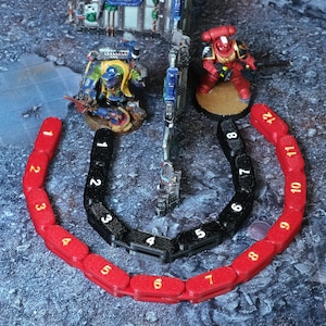 Può includere: Una scena di gioco da tavolo con miniature e accessori. Due indicatori segmentati, uno rosso e uno nero, numerati da 1 a 12, sono in primo piano. Una miniatura rossa e nera è sullo sfondo.