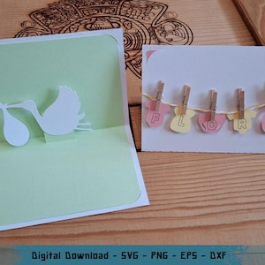 May include: A handmade baby shower card with a pop-up stork carrying a baby. The card is light green and white. Another card displays the name "FLORA" spelled out with letters attached to baby-themed cutouts, held by clothespins on a string.