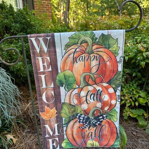 May include: A rectangular garden flag featuring a vertical brown wood-grain panel on the left with the word WELCOME printed in white. The right side displays three stacked orange pumpkins with green leaves, a black and white buffalo check bow, and the text happy fall y'all.