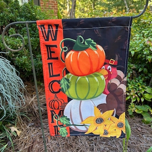 May include: A rectangular garden flag featuring a vertical orange side panel with the black text WELCOME. The main black section displays a stack of three pumpkins in orange, green, and white, a turkey with brown feathers, and yellow flowers.