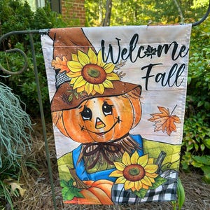 May include: A rectangular garden flag featuring a scarecrow with a pumpkin head, wearing a brown hat adorned with a yellow sunflower. The text Welcome Fall is printed in black script. The design includes orange maple leaves and sunflowers.