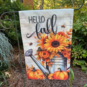 May include: A rectangular garden flag featuring the text Hello fall in black script and block lettering. The design displays a silver watering can filled with orange sunflowers and autumn leaves, accented by two orange pumpkins and bees.