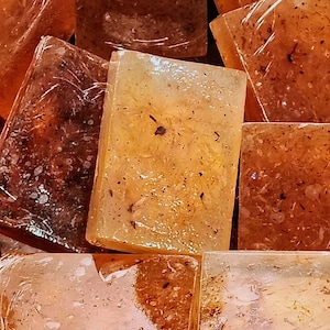 May include: Close-up of several rectangular bars of soap. The soaps are various shades of amber and brown, some are wrapped in clear plastic. The soaps have visible flecks of what appears to be herbs or botanicals. The soaps are arranged closely together.