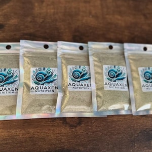 May include: Five foil pouches filled with a light green powder, arranged in a row. Each pouch has a label with a blue and white snail design and the text "AQUAXEN NUTRITION". The pouches are on a dark wood surface.
