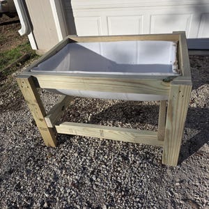 Op de afbeelding: Een verhoogde plantenbak van hout met een witte plastic voering. Het frame is gemaakt van onbewerkt hout en heeft een rechthoekige vorm. De bak is ontworpen om op de grond te staan en is geschikt voor het kweken van planten.