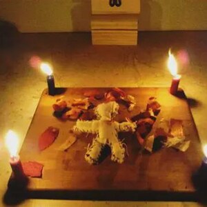 May include: A voodoo doll lies on a wooden surface, surrounded by four lit candles. The doll is made of white fabric with stitched details. Dried leaves and red paper pieces are scattered around the doll, creating a ritualistic scene.