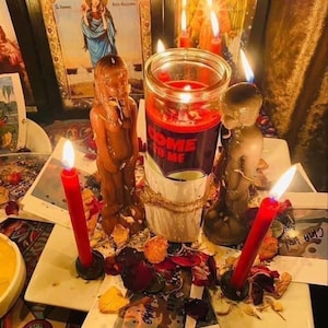 Puede incluir: Una vela central en frasco de vidrio con una etiqueta roja que dice COME TO ME, flanqueada por dos figuras de cera con forma humana y dos velas rojas largas. El conjunto se asienta sobre un plato cuadrado blanco con pétalos de flores secas.