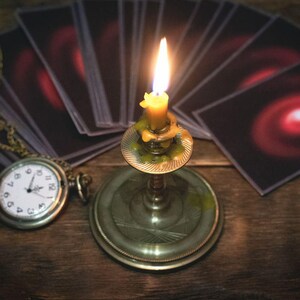 Puede incluir: Una vela encendida en un candelero de latón, un reloj de bolsillo y una baraja de cartas de tarot con diseños de espirales rojas sobre una superficie de madera. La escena evoca misterio y adivinación.