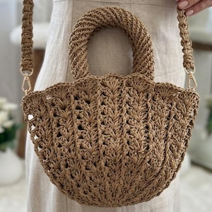 May include: A woven brown handbag with a rounded handle and a shoulder strap. The bag has a scalloped edge and is held by a person wearing a beige dress. The bag is made of a natural fiber material.