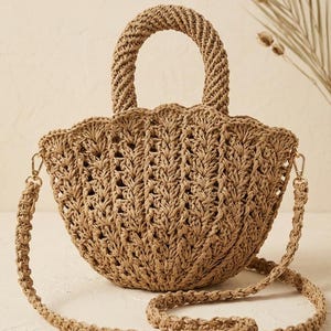 Peut inclure: Un sac à main tissé couleur beige en forme de coquillage avec un design texturé et festonné. Le sac comprend deux poignées supérieures arrondies et robustes ainsi qu'une longue bandoulière tressée amovible fixée par des fermoirs dorés.