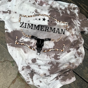 May include: A tie-dye t-shirt with a brown and white pattern. The shirt features the name "ZIMMERMAN" in black letters, a black bull skull, and leopard print accents. The design includes stars and a decorative border.