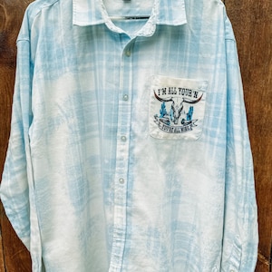 May include: A light blue plaid button-up shirt with a white bleached effect. The shirt has a pocket with a graphic of a skull and the text "I'm all your'n you're all mine".