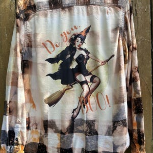May include: A long-sleeve button-down shirt with a bleached plaid pattern in shades of black, white, and orange. The back features a graphic of a pin-up style witch flying on a broomstick with the orange text Do you. BOO! printed around the figure.