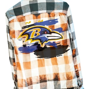 May include: A white, orange, and black plaid flannel shirt with a Baltimore Ravens logo on the back. The logo is a purple and black raven with the letter "B" on its chest.