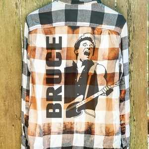 May include: A white and black plaid flannel shirt with a brown bleach design. The design features a silhouette of a person playing a guitar with the word "BRUCE" printed vertically on the left side.