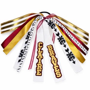 May include: A hair accessory featuring multiple ribbons in gold, maroon, and white. The ribbons include text reading CLAIRE and BRAVES, along with XC symbols and arrow graphics. The ribbons are attached to a black elastic hair tie.