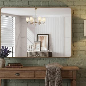 May include: A rectangular gold-framed mirror hangs above a wooden console table. A macrame wall hanging and a vase of purple flowers decorate the green brick wall. A wooden floor lamp with a beige lampshade stands to the right.
