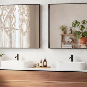 May include: A modern bathroom vanity with two square mirrors framed in black. The vanity has a wooden cabinet with two white rectangular sinks and black faucets. Decorative items include plants, soaps, and woven wall hangings.