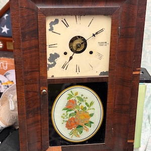 May include: Antique wooden clock with a rectangular frame. The clock face is off-white with black Roman numerals and hands. Below the clock face is a circular floral design with orange and yellow flowers. The clock has a dark brown finish.