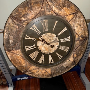 May include: Large, round wall clock with a distressed, textured wooden frame. The clock face is dark with Roman numerals and ornate hands. The clock's outer frame has a decorative, bark-like appearance.