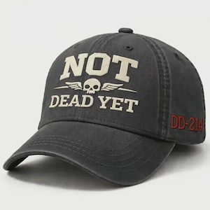 Op de afbeelding: Donkergrijze baseballcap met de witte tekst "NOT DEAD YET" en een schedel-met-vleugels-afbeelding. De tekst "DD-214" is in het rood op de zijkant geborduurd. Een casual accessoire.