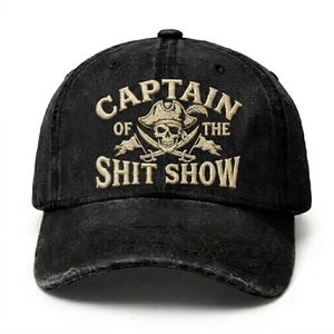 Op de afbeelding: Zwarte baseballcap met de geborduurde tekst "CAPTAIN OF THE SHIT SHOW" in beige. Het ontwerp bevat een schedel met een piratenhoed, gekruiste zwaarden en veren. De cap heeft een gebogen klep en een verstelbare band.