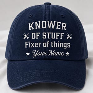 May include: A navy blue baseball cap featuring white embroidered text that reads KNOWER OF STUFF, Fixer of things, and Your Name. The design includes two crossed wrench icons and two stars flanking the bottom line of text on the front panel.
