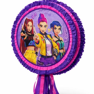May include: A circular piñata with a purple and pink frilled edge, featuring an image of three animated women. The women are dressed in vibrant outfits. Purple and pink ribbons dangle from the base. A festive party decoration.