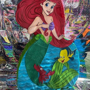 May include: A colorful, cartoon-style cutout of Ariel from The Little Mermaid. Ariel has red hair, a purple seashell top, and a green tail. Flounder and Sebastian are also featured. The background is a shiny, iridescent material.