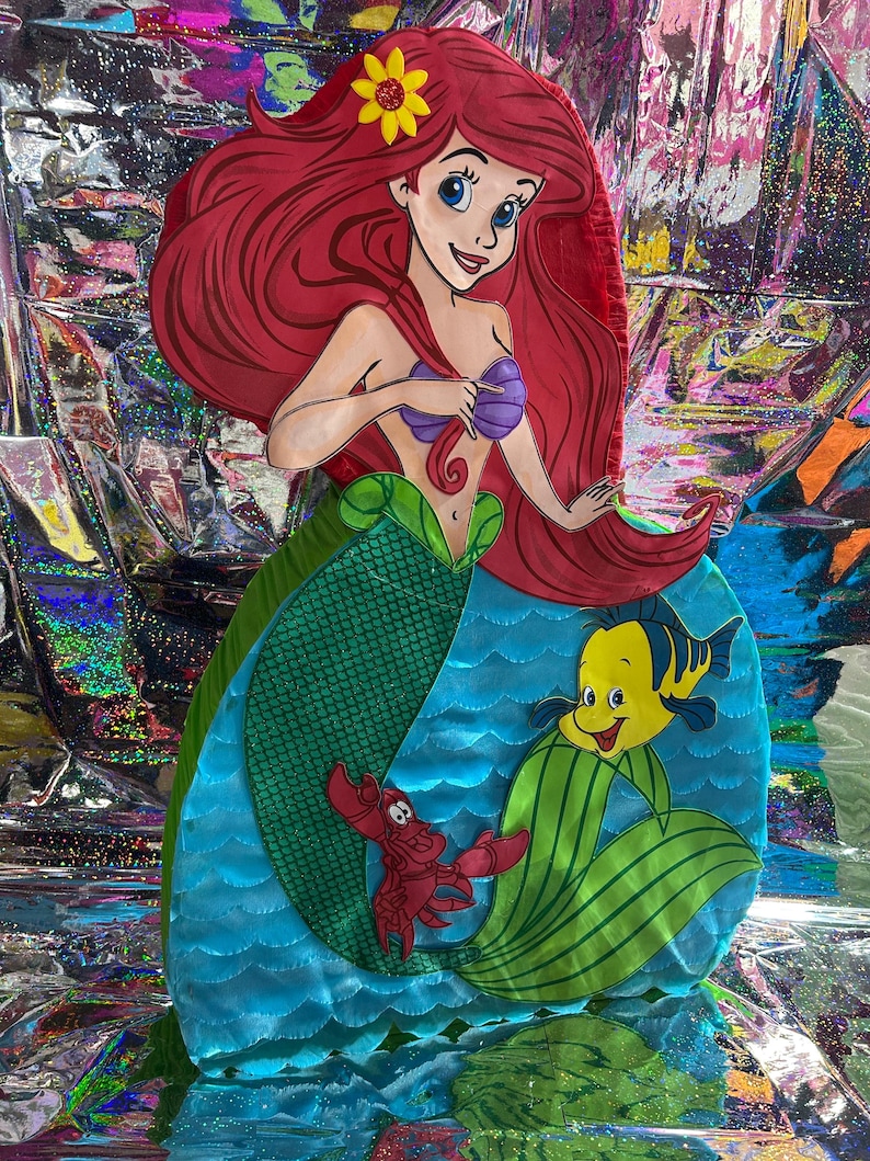 May include: A colorful, cartoon-style cutout of Ariel from The Little Mermaid. Ariel has red hair, a purple seashell top, and a green tail. Flounder and Sebastian are also featured. The background is a shiny, iridescent material.